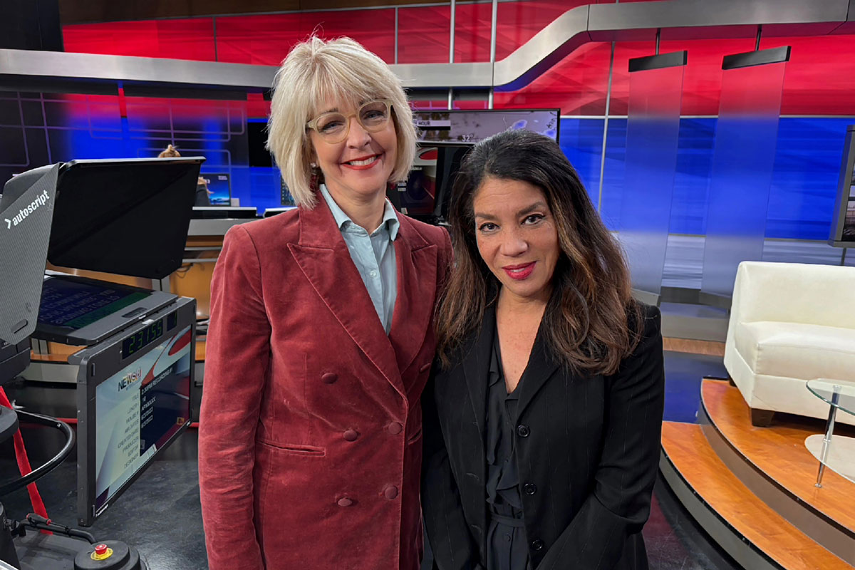 Two women pose and smile together on a news set
